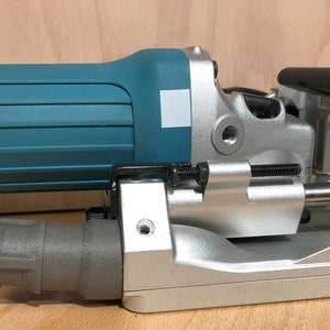 Könnte beinhalten: Ein türkis-schwarzes Elektrowerkzeug mit silberfarbenem Metallsockel. Das Werkzeug hat einen zylindrischen Körper und eine rechteckige Basis mit einem schwarzen Griff. Das Werkzeug ist für die Holzbearbeitung und den Bau bestimmt.