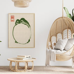 May include: A green frog illustration in a circle with the Japanese character "圓" in red above it, framed and hanging on a white wall. A wicker hanging chair with a white throw blanket and two white pillows is in the foreground. A wicker coffee table with books on top is in the middle of the room.