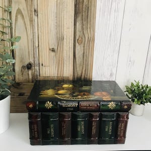 May include: A decorative box designed to resemble a stack of books. The box features a dark, glossy top with a still-life painting. The sides are designed to look like book spines, with titles including "Gone With The Wind" and "Egmont 1788".