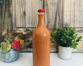 Puede incluir: Una botella de cerámica de color terracota con un tapón de madera y rojo. La botella está sobre una superficie de madera.