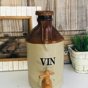 Puede incluir: Una jarra de vino de cerámica con un esmalte marrón y beige. La jarra tiene un grifo de madera y la palabra "VIN" escrita en la parte delantera.