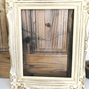 May include: A rectangular picture frame featuring an ornate, distressed finish with a combination of cream and metallic gold paint. The frame has a raised, carved border design and includes a thin wire attached to the back for hanging purposes.