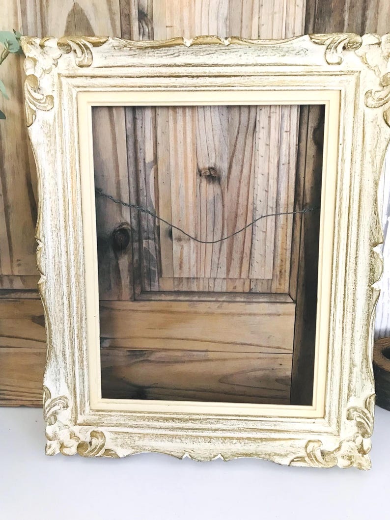 May include: A rectangular picture frame featuring an ornate, distressed finish with a combination of cream and metallic gold paint. The frame has a raised, carved border design and includes a thin wire attached to the back for hanging purposes.