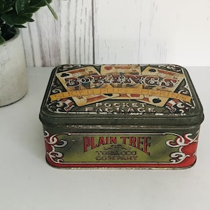 Może przedstawiać: Winietkowy, zielony metalowy pojemnik z czerwono-biało-żółtym wzorem. Pojemnik zawiera karty do gry i tekst "Bokings Sliced Plug Smoking Pocket Package" oraz "Plain Tree Tobacco Company".