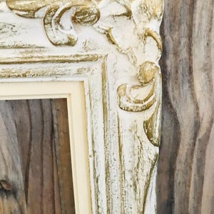May include: A rectangular picture frame featuring an ornate, raised relief pattern along the edges. The frame has a distressed finish with a base of off-white paint accented by gold-toned highlights on the decorative scrollwork and corner details.