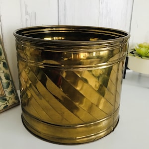 May include: A gold metal planter with a decorative, diagonal pattern. The planter is round and has a smooth, shiny finish.