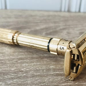 Op de afbeelding: Een goudkleurig veiligheidsscheermes met een getextureerde handgreep en verstelbare instellingen. Het scheermes ligt op een houten oppervlak. De instelknop is gemarkeerd met het nummer 5.