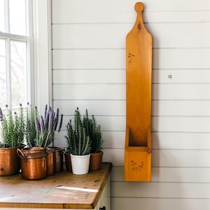 May include: A tall, vertical wooden wall pocket with a light brown finish and a rounded top handle. The piece features small, dark floral engravings on the upper and lower sections, providing a functional storage space for small items.