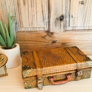 May include: A rectangular suitcase with a textured brown faux-alligator skin pattern, featuring metallic corner reinforcements, two vertical straps with buckles, and a sturdy orange handle. The case has a structured design with visible rivets.