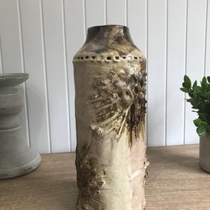May include: A tall, ceramic vase with a textured, brown and white glaze. The vase has a cylindrical shape and a slightly flared opening. The glaze has a rough, uneven texture, creating a unique and rustic look.