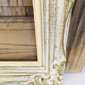 May include: A rectangular picture frame featuring a distressed finish with layers of white and gold paint. The frame has ornate, carved detailing along the edges and a smooth, cream-coloured inner border surrounding the central rectangular opening.