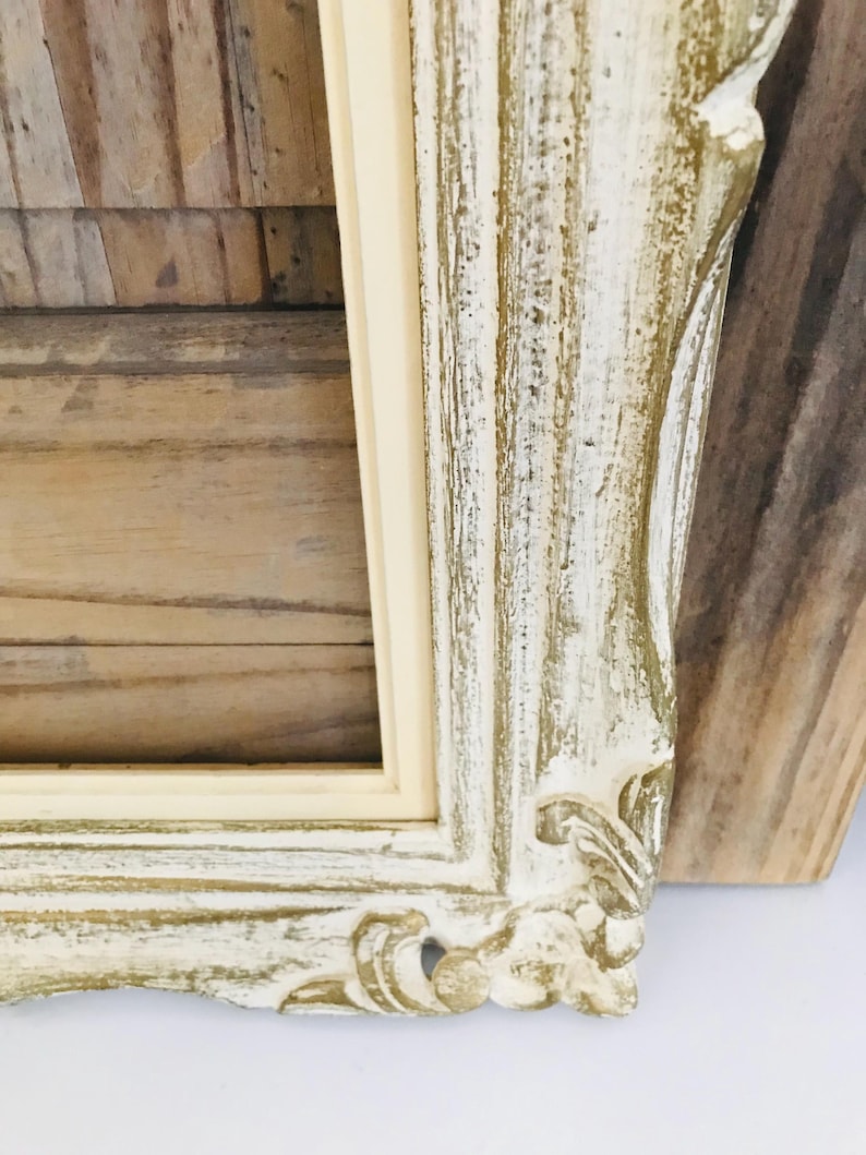 May include: A rectangular picture frame featuring a distressed finish with layers of white and gold paint. The frame has ornate, carved detailing along the edges and a smooth, cream-coloured inner border surrounding the central rectangular opening.