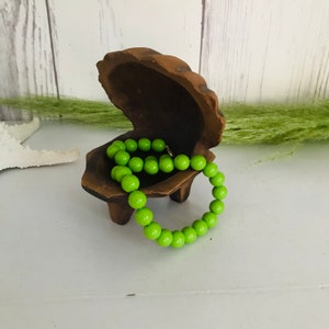 May include: A bright green beaded necklace rests inside a dark brown, wooden clam shell-shaped jewelry box. The box is open, revealing the necklace's vibrant color against the wood. The necklace is made of round beads.