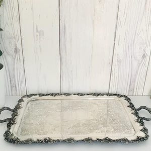 Könnte beinhalten: Ein silbernes Serviertablett mit einem verzierten, geprägten Design und zwei Griffen. Das Tablett ist rechteckig und hat einen leicht erhöhten Rand.