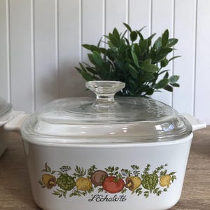 May include: White ceramic casserole dish with a glass lid. The dish features a green, yellow and red floral design with the words "L'Echalote" printed on the front.