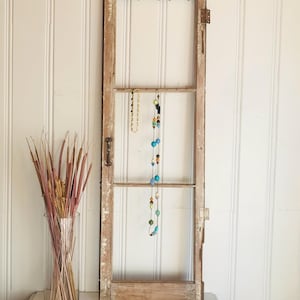 May include: A vintage wooden window frame, repurposed as a jewellery organiser. Two necklaces hang from the frame, one gold-toned and the other with colourful beads. A clear glass vase with dried plants sits to the left, against a white panelled wall.