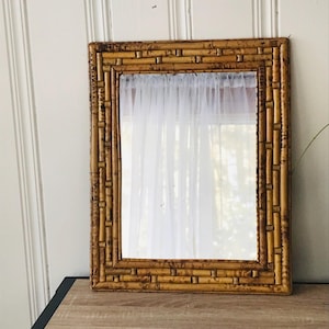 May include: Rectangular mirror with a bamboo frame. The frame is made of multiple bamboo stalks, creating a textured, natural look. The mirror reflects a sheer white curtain. The mirror is a decorative item.