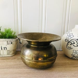 Puede incluir: Un recipiente de latón vintage con una parte superior ancha y poco profunda y una base redondeada. El latón tiene una pátina cálida y envejecida. El recipiente se exhibe sobre una superficie de madera, con una pequeña planta en maceta y un jarrón decorativo en el fondo.