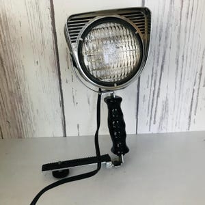 May include: A vintage chrome spotlight with a black handle and a clamp. The spotlight has a large, round, clear glass lens with a grid pattern.