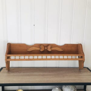 May include: A vintage wooden shelf with a decorative bow carving. The shelf has a lower rail with small spindles and is made of light brown wood. It is designed to be mounted on a wall and is suitable for displaying small items.