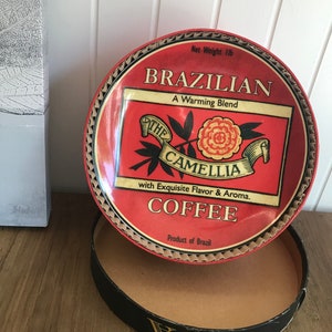 以下が含まれることがあります： ブラジルのコーヒーの赤い金色の缶。缶には「ブラジリアン、ウォーミングブレンド、ザ・カメリア、エキゾチックなフレーバーとアロマ、コーヒー、ブラジル産」と書いてあります。缶は開いていて、蓋は缶の上に置かれています。蓋は赤と金のデザインで、花と葉が描かれています。