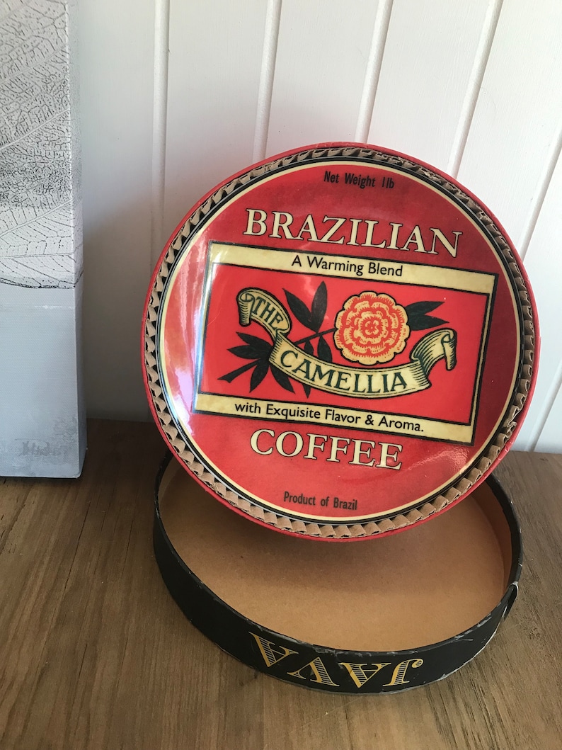 May include: A red and gold tin with a label that reads "Brazilian, A Warming Blend, The Camellia, with Exquisite Flavor & Aroma, Coffee, Product of Brazil". The tin is open and the lid is sitting on top of the tin. The lid has a red and gold design with a flower and leaves.
