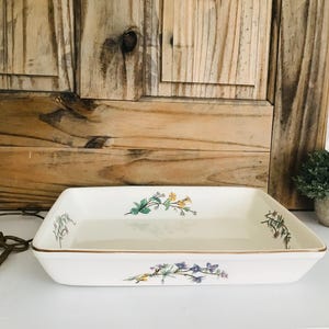Puede incluir: Una fuente rectangular de cerámica blanquecina con diseños florales en verde, amarillo y morado. La fuente tiene un borde dorado y está colocada sobre un fondo de madera. También se ven una pequeña planta en maceta y llaves antiguas.