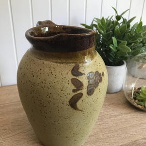 Op de afbeelding: Een bespikkelde, keramische kan met een afgeronde body en een donkerbruine rand en handvat. De kan heeft een lichtgroene basiskleur met bruine bloemmotieven. De kan staat op een houten oppervlak.