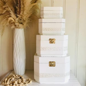 Peut inclure: Une pile de quatre boîtes octogonales blanches avec une bordure en relief et des fermoirs en métal doré sur les deux plus grandes boîtes. Les boîtes sont disposées verticalement, de la plus grande à la plus petite, avec une finition mate.