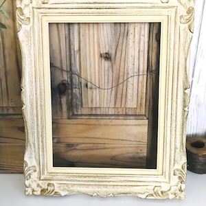 May include: A rectangular picture frame featuring an ornate, carved border with a distressed finish in cream and gold tones. The frame includes a smooth, light-coloured inner moulding and a thin metal wire attached to the back for hanging purposes.