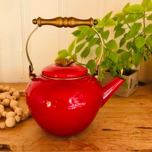 May include: A bright red apple-shaped tea kettle with a glossy finish. The kettle features a gold-toned metal handle and a matching gold-toned leaf-shaped knob on the lid. It has a curved spout and a rounded body, designed for stovetop use.