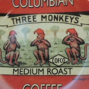 May include: A red and white coffee can with a picture of three monkeys wearing red hats. The monkeys are covering their eyes, ears, and mouth. The can says "Columbian Three Monkeys Medium Roast Coffee" and "COFCO".