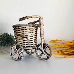 May include: A wicker basket on wheels shaped like a bicycle. The basket is light brown and has a white wash finish. The wheels are black with a white wash finish.