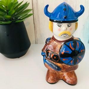 Könnte beinhalten: Eine Keramikfigur eines Wikingerkriegers mit einem blauen Helm mit Hörnern, einer braunen Tunika und einem blauen und schwarzen Schild.