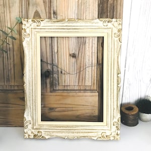 May include: A rectangular picture frame featuring an ornate, carved border with a distressed cream and gold finish. The frame has a smooth inner edge and a thin wire attached to the back for hanging, showcasing a classic, decorative design.