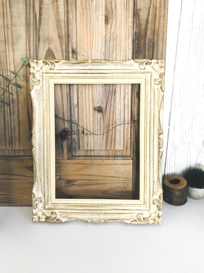 May include: A rectangular picture frame featuring an ornate, carved border with a distressed cream and gold finish. The frame has a smooth inner edge and a thin wire attached to the back for hanging, showcasing a classic, decorative design.