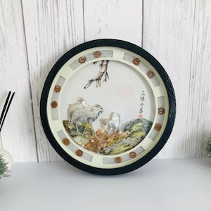 Peut inclure: Une décoration murale ronde encadrée de noir avec un fond blanc et une scène 3D de moutons blancs dans un paysage montagneux. La scène est faite de coquillages et d'autres matériaux naturels. Le cadre a une bordure décorative de coquillages bruns et blancs.