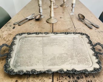 May include: A rectangular silver-toned serving tray featuring an ornate embossed border with grape and vine motifs, two side handles, and a central engraved pattern. The tray is positioned on a wooden surface with silver cutlery and candle holders.