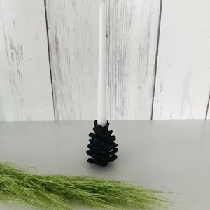 May include: A black pine cone candle holder with a white candle. The candle holder is made of metal and has a rustic look.