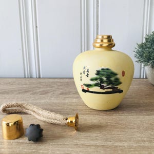 May include: A cream-coloured oil lamp with a gold-coloured top and a painted tree design. The lamp includes a gold-coloured cap, a black star-shaped piece, and a wick with a gold-coloured end. The lamp is approximately 15 cm tall.