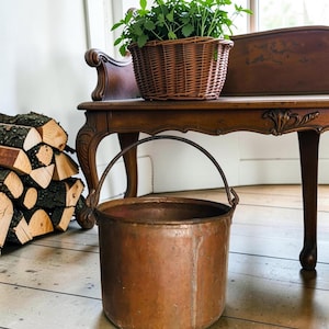 May include: A cylindrical copper bucket with a weathered patina and a dark metal swing handle. The bucket features a smooth, rounded body and a visible seam along the side, showcasing a metallic texture and warm, reddish-brown tones throughout.