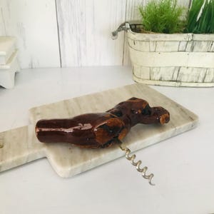 May include: A brown ceramic corkscrew shaped like a bird with a metal corkscrew attached to the bottom. The corkscrew is sitting on a white marble cutting board.