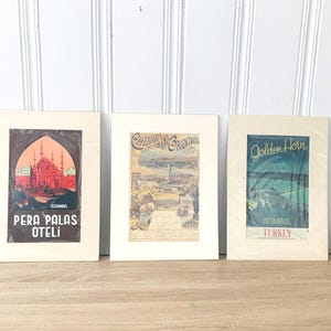 May include: Three framed vintage travel posters of Istanbul, Turkey. The posters have cream-coloured mounts and depict scenes of the city, including the Pera Palas Oteli, the Golden Horn, and a coastal view. The posters are in protective plastic.