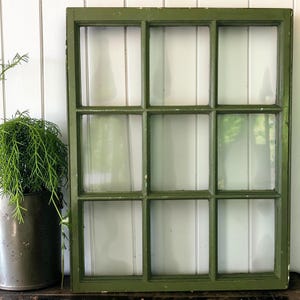 May include: A rectangular window frame painted in a matte olive green finish, featuring a grid of nine individual glass panes arranged in three rows and three columns. The wooden frame shows slight signs of wear along the edges and corners.