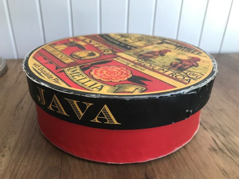 May include: A red and black round tin box with the word "JAVA" in gold lettering. The lid features a vintage design with the words "Amelia Blend" and "Medium Roast" in a yellow banner.