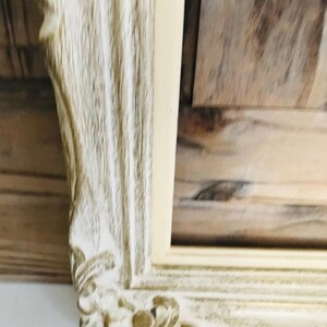 May include: A rectangular picture frame featuring an ornate, carved border with a distressed finish. The frame displays a combination of off-white and light brown wood tones, highlighting the intricate scrollwork details at the corner.