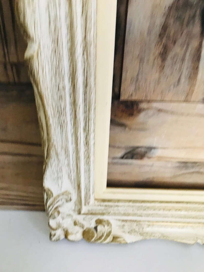 May include: A rectangular picture frame featuring an ornate, carved border with a distressed finish. The frame displays a combination of off-white and light brown wood tones, highlighting the intricate scrollwork details at the corner.