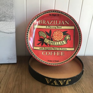 May include: A red and gold tin can with a white label that says "Brazilian Coffee, A Warming Blend, The Camellia, with Exquisite Flavor & Aroma. Product of Brazil". The tin can has a black and gold lid with the word "VAVI" printed on it.