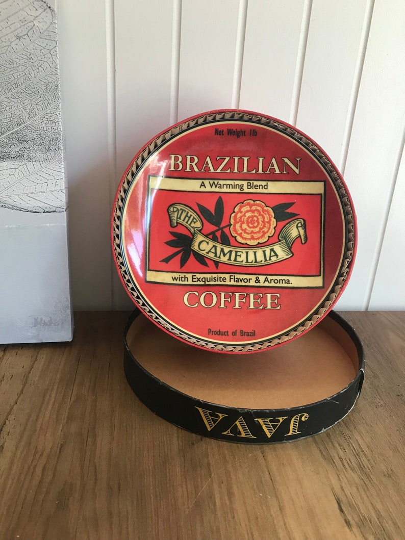May include: A red and gold tin can with a white label that says "Brazilian Coffee, A Warming Blend, The Camellia, with Exquisite Flavor & Aroma. Product of Brazil". The tin can has a black and gold lid with the word "VAVI" printed on it.
