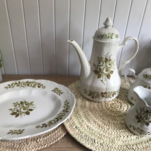 Könnte beinhalten: Ein weißes Porzellan-Tee-Set mit floralem Design. Das Set beinhaltet eine Teekanne, eine Milchkännchen, eine Zuckerdose und eine Servierplatte. Das Blumenmuster zeigt grüne Blätter und weiße Rosen.
